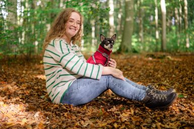Frau sitzt im Wald auf Laub mit einem kleinen Hund in rotem Pullover.