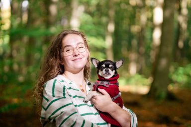Eine Frau im Wald hält einen kleinen Hund in einem roten Outfit.